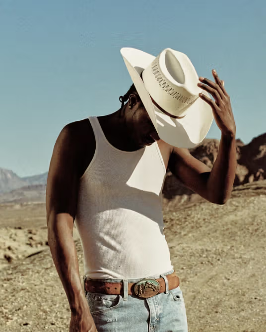 A person wearing a white tank top, jeans, and a white cowboy hat stands outdoors in a desert landscape, holding the brim of the hat and looking down.