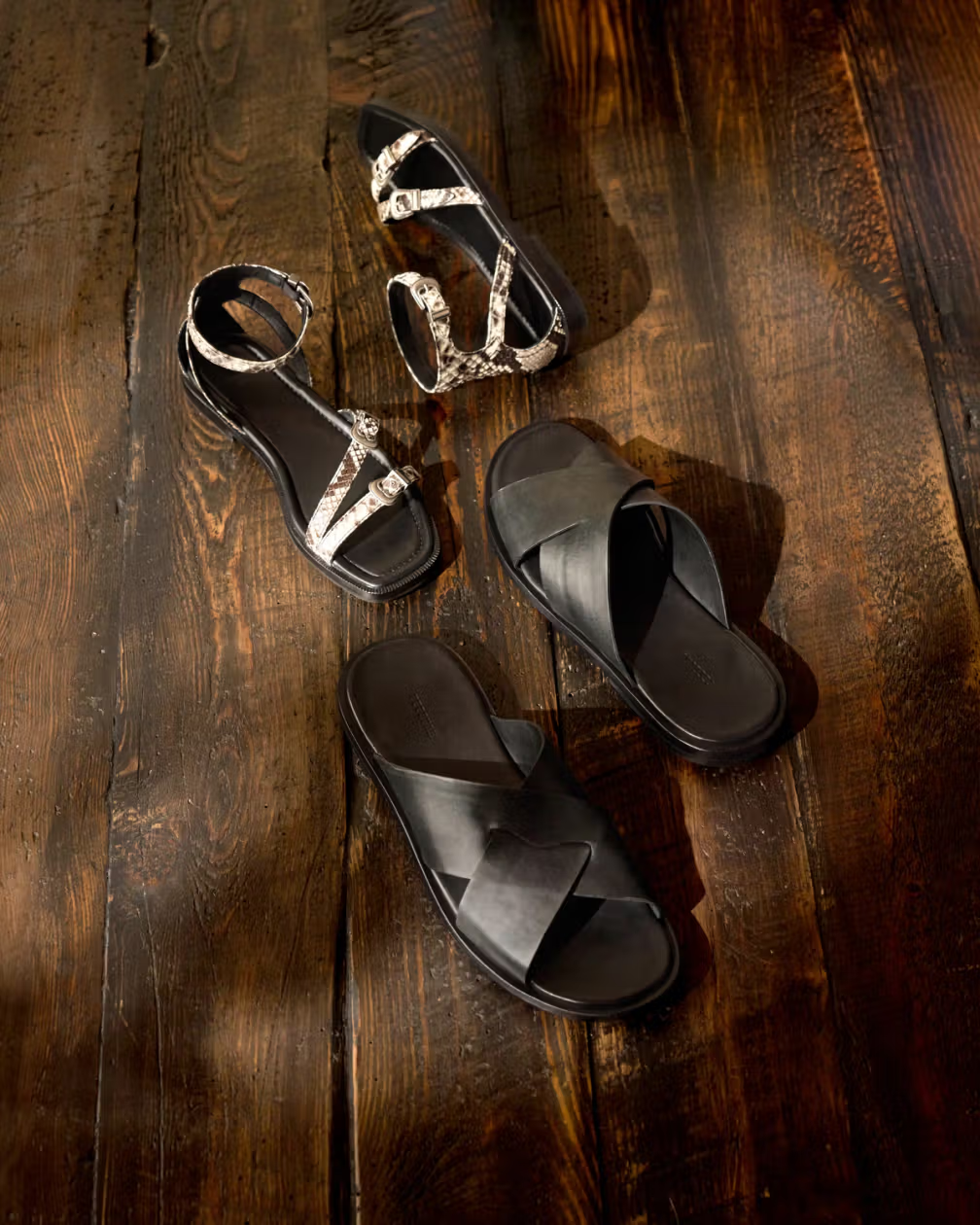 Four pairs of sandals and slides are arranged on a dark wooden surface.