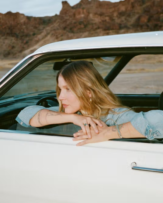 A person with long blond hair leans out of the window of a white car, resting their arms on the door, with a desert landscape in the background.