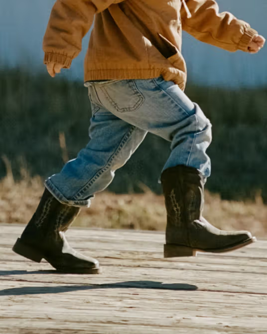 A child wearing a brown jacket, blue jeans, and black cowboy boots walks outdoors on a wooden surface.