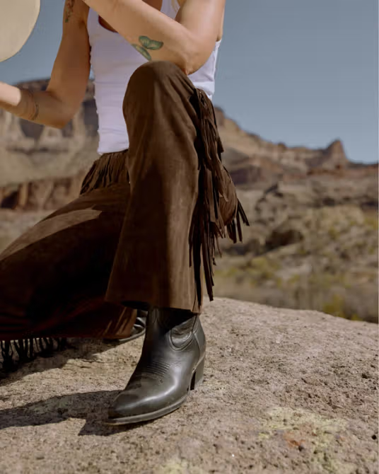 A person in a white tank top and brown fringe pants sits on a rock outdoors, wearing black heeled boots and holding a hat; desert landscape in the background.