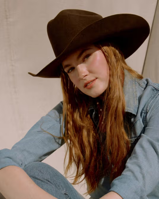 A woman with long red hair wearing a wide-brimmed brown hat and a denim shirt sits outdoors, looking toward the camera.