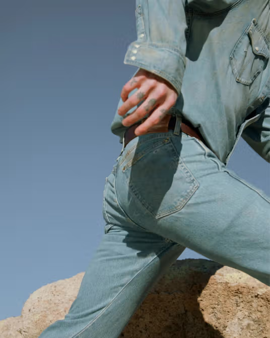 Person wearing light blue denim shirt and jeans, climbing over a rock outdoors; only torso, arm, and legs visible against a clear sky.
